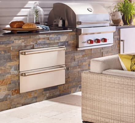 Outdoor kitchen featuring a built-in Wolf appliance stainless steel grill, stone façade, side drawers, countertop with bread and plates, and a wicker sofa in the foreground.