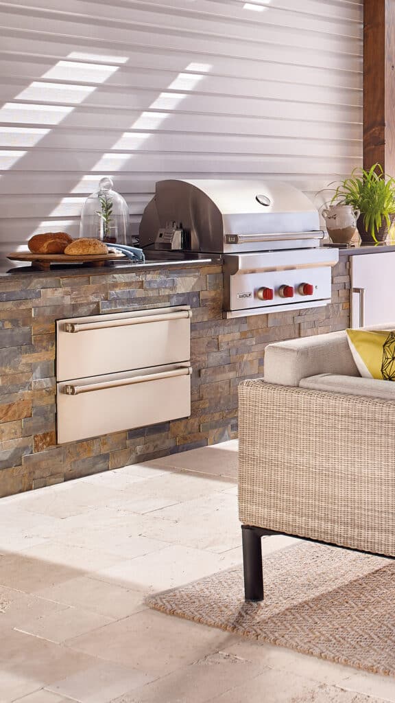 Outdoor kitchen featuring a built-in Wolf appliance stainless steel grill, stone façade, side drawers, countertop with bread and plates, and a wicker sofa in the foreground.
