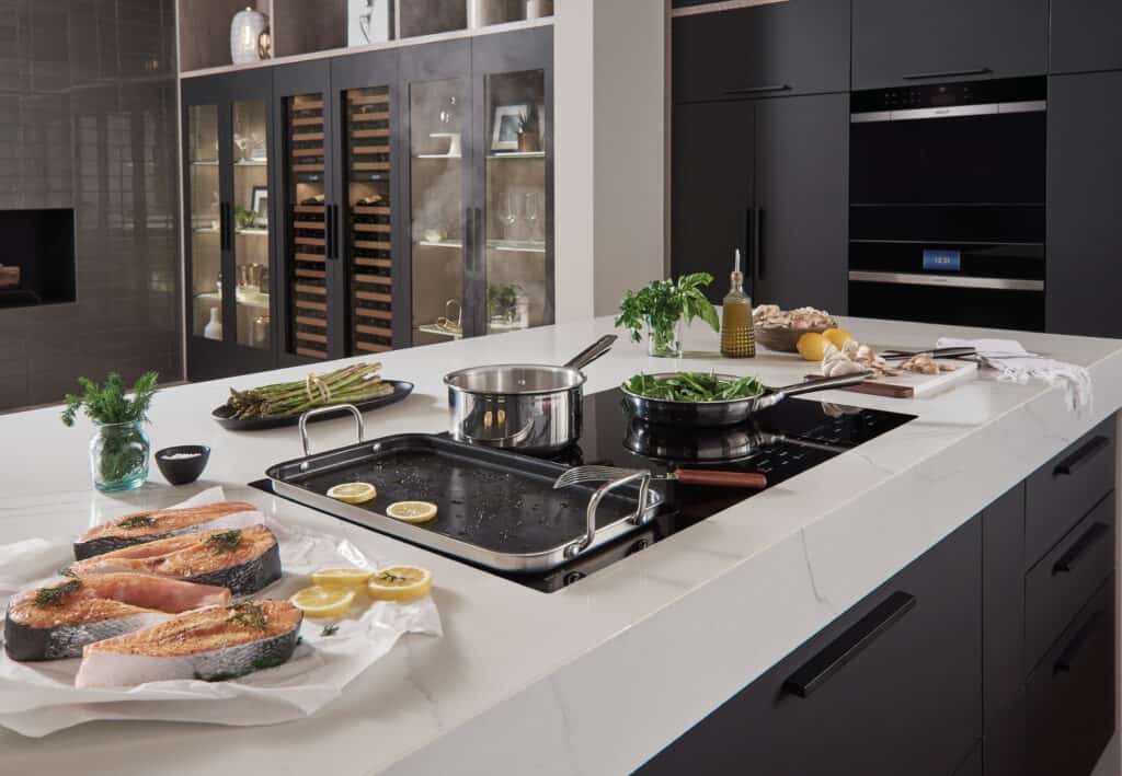 A modern kitchen countertop with raw salmon, lemon slices, vegetables, pots, and pans prepared for cooking, highlighted by contemporary black cabinets and luxury kitchen appliances in the background.
