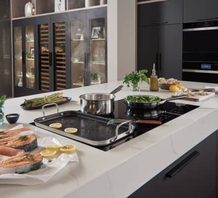A modern kitchen countertop with raw salmon, lemon slices, vegetables, pots, and pans prepared for cooking, highlighted by contemporary black cabinets and luxury kitchen appliances in the background.