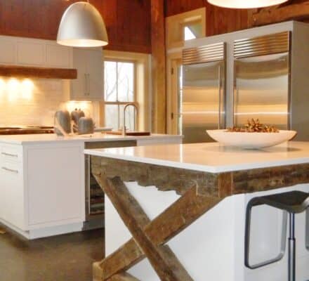 Modern kitchen showroom with white island, exposed wooden beams, pendant lights, stainless steel appliances including luxury Wolf appliance, and wood-paneled walls and ceiling. Two metal bar stools complete the stylish space.