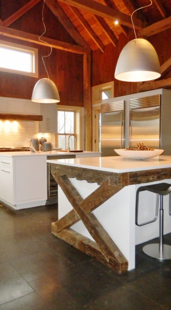 Modern kitchen showroom with white island, exposed wooden beams, pendant lights, stainless steel appliances including luxury Wolf appliance, and wood-paneled walls and ceiling. Two metal bar stools complete the stylish space.