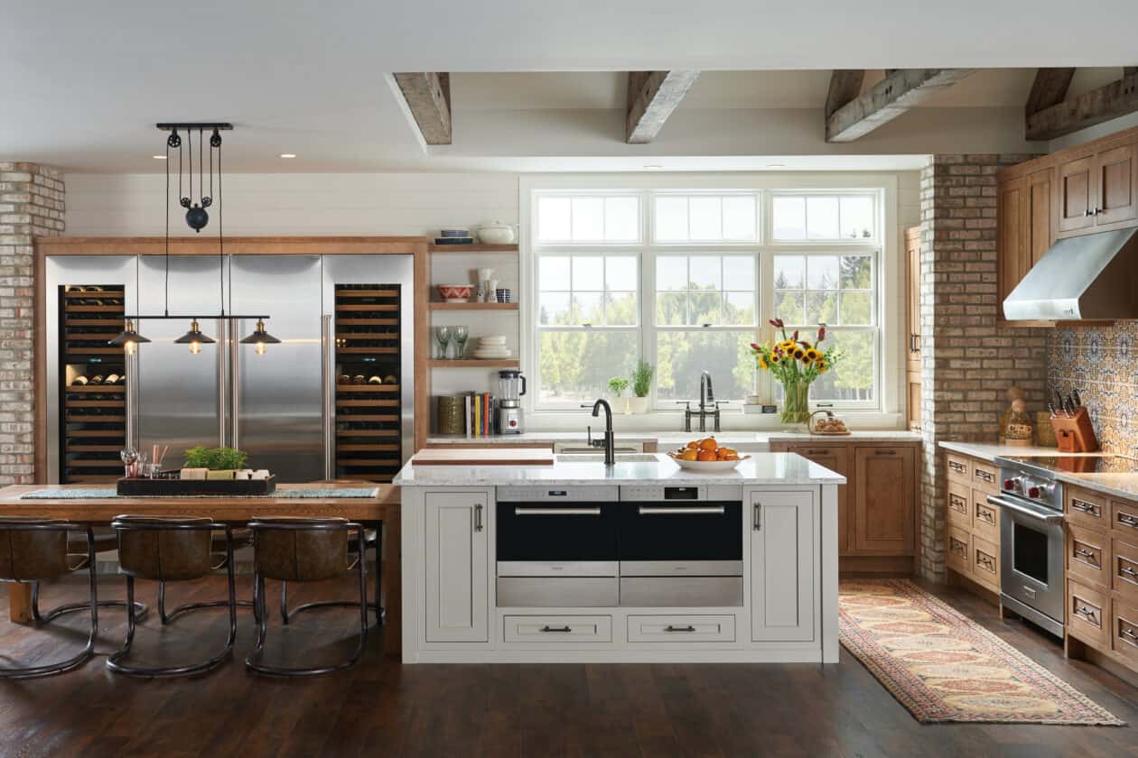 Modern kitchen showroom with brick walls, stainless steel luxury kitchen appliances, a spacious island with a sink, wooden cabinets, dining table with chairs, and large windows filling the space with natural light.