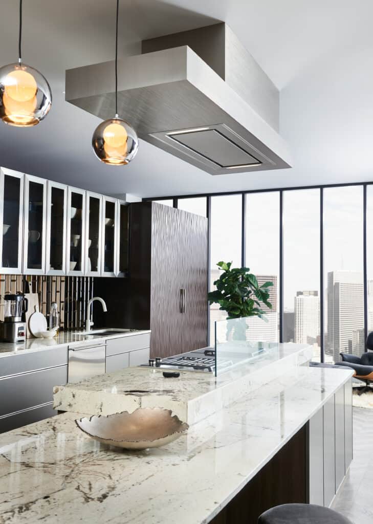 Modern kitchen with marble island countertop, luxury kitchen appliances, hanging pendant lights, a ventilation hood, and floor-to-ceiling windows showcasing city buildings in the background—perfect inspiration for any appliance showroom.