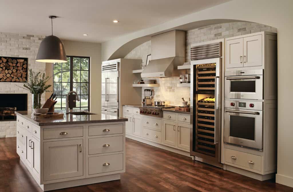 Modern kitchen with white cabinets, stainless steel appliances—including a Wolf appliance—wine fridge, large island with sink, and hardwood floors. Features a window and built-in shelves with stacked firewood. Perfect for any luxury kitchen appliance enthusiast.