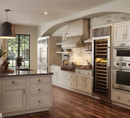 Modern kitchen with white cabinets, stainless steel appliances—including a Wolf appliance—wine fridge, large island with sink, and hardwood floors. Features a window and built-in shelves with stacked firewood. Perfect for any luxury kitchen appliance enthusiast.
