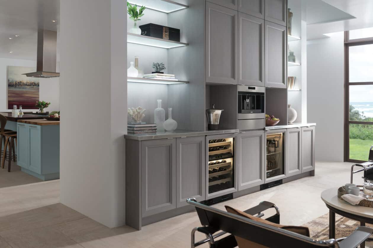 Modern kitchen with built-in gray cabinets, open shelves with decor, integrated wine cooler, coffee machine, and large windows providing natural light—showcasing the elegance of a luxury kitchen appliance from a top appliance showroom.