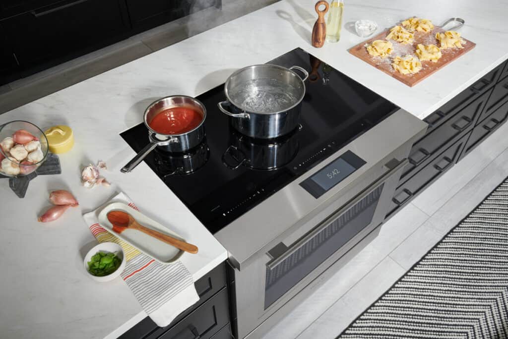 A modern kitchen showroom with an induction cooktop; one pot with tomato sauce, another with boiling water, and cooking ingredients, including pasta, on the counter—showcasing the latest in luxury kitchen appliance design.