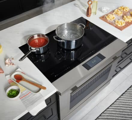 A modern kitchen showroom with an induction cooktop; one pot with tomato sauce, another with boiling water, and cooking ingredients, including pasta, on the counter—showcasing the latest in luxury kitchen appliance design.
