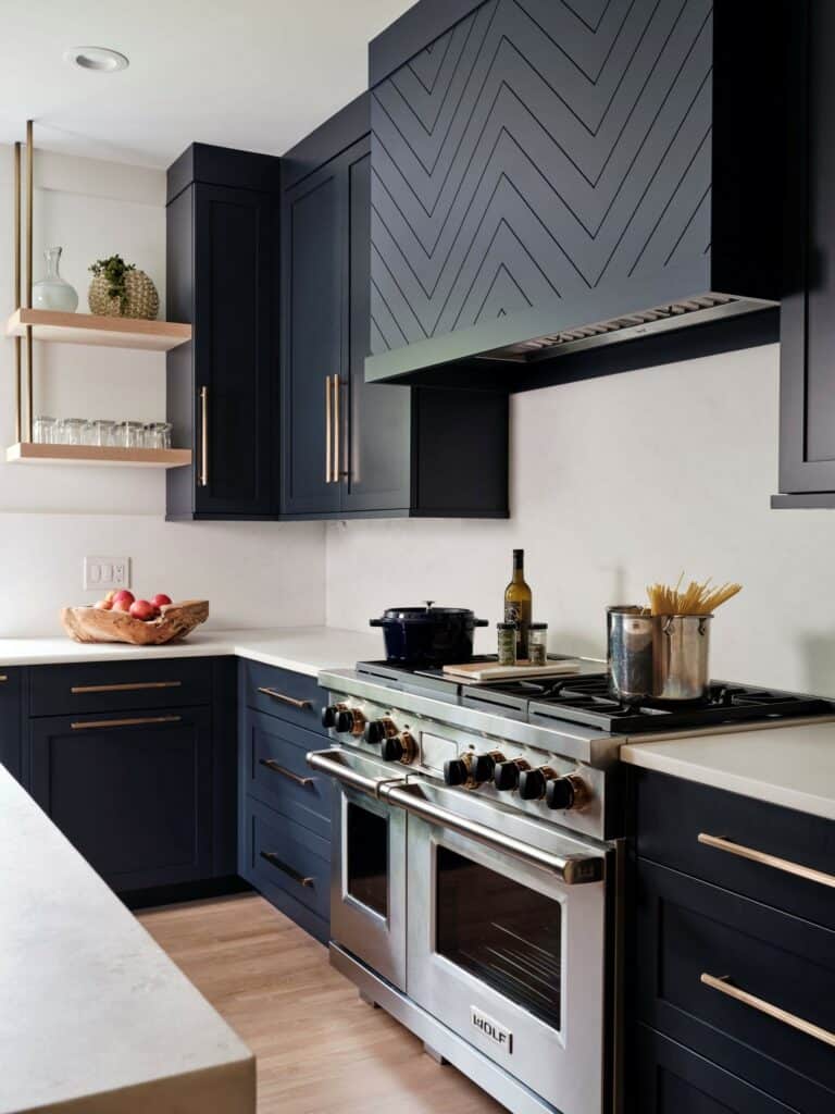 Modern kitchen with navy cabinets, gold hardware, a stainless steel stove, sleek Wolf appliance, cookware, open wood shelves, and a bowl of apples on a light countertop.