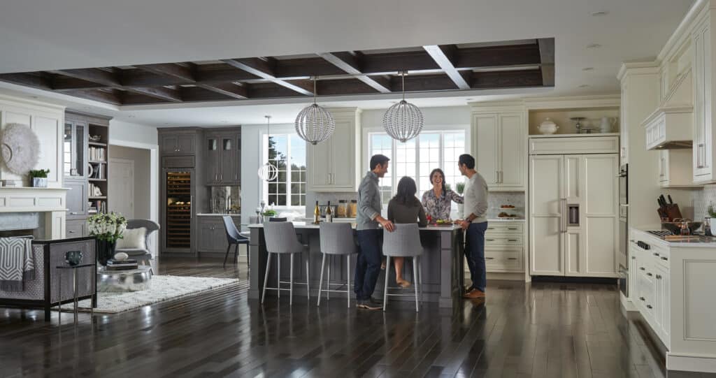 Four people stand and converse around a kitchen island in a modern, spacious kitchen showroom with white cabinetry, dark wood floors, pendant lighting, and sleek Sub Zero appliances.