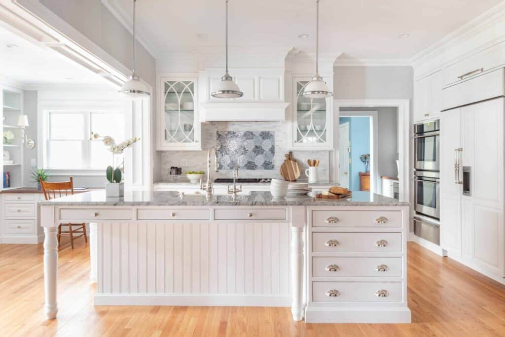 Bright, spacious kitchen with white cabinetry, marble island, stainless steel appliances—including a Sub Zero appliance—wooden floors, and pendant lights above the island, perfect for a kitchen showroom display.