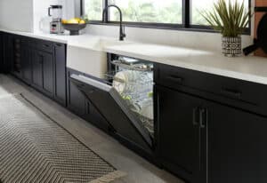 A modern kitchen with black cabinets and a white countertop features an open luxury kitchen appliance dishwasher filled with dishes beneath a large window.