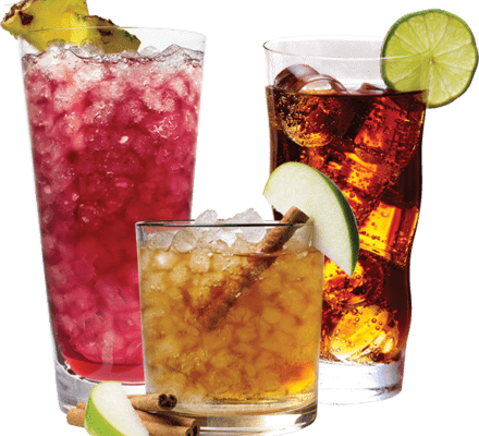 Three iced drinks in a kitchen showroom: a pink cocktail with pineapple, a brown cocktail with apple slice and cinnamon stick, and a dark soda with a lime wheel. Perfect sips while browsing the latest Wolf appliance displays.