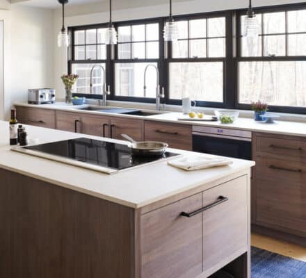 Modern kitchen with a large island, Wolf Induction Cooktop, wood cabinetry, pendant lights, and large windows providing natural light. Items and utensils are neatly arranged on the counters.