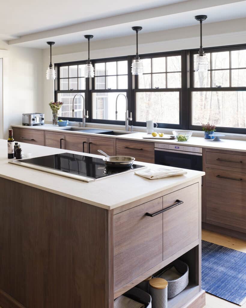 Modern kitchen with a large island, Wolf Induction Cooktop, wood cabinetry, pendant lights, and large windows providing natural light. Items and utensils are neatly arranged on the counters.