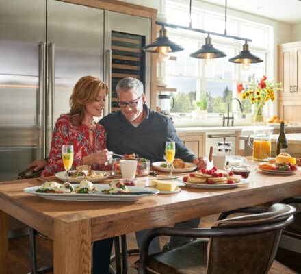 A man and woman sit together at a kitchen table set with breakfast foods, smiling at a phone. The bright space features modern decor and luxury kitchen appliances, with large windows flooding the stylish kitchen showroom with light.