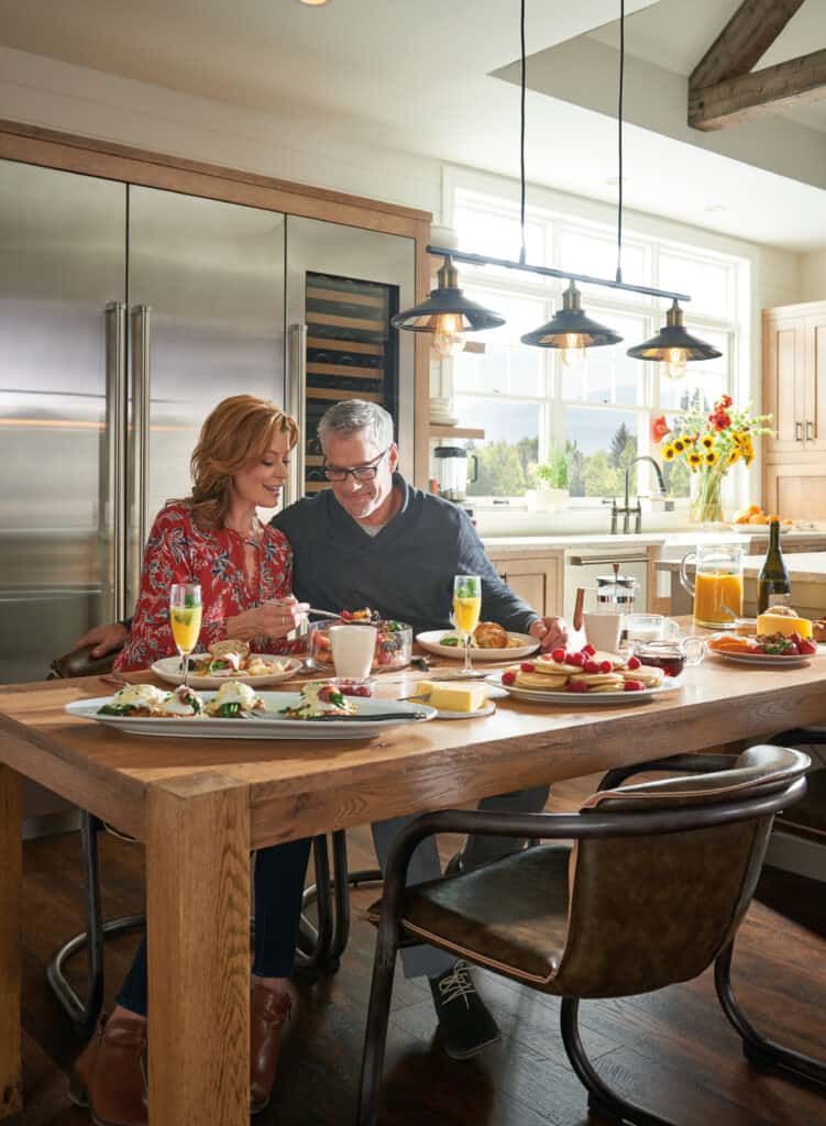 A man and woman sit together at a kitchen table set with breakfast foods, smiling at a phone. The bright space features modern decor and luxury kitchen appliances, with large windows flooding the stylish kitchen showroom with light.