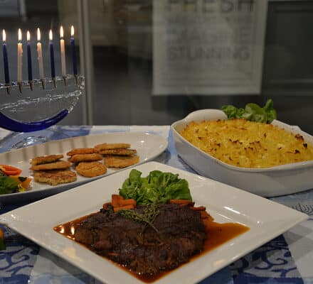 A Hanukkah table setting with a lit menorah, latkes, kugel, and brisket with greens on a blue-patterned tablecloth, perfect for showcasing in an elegant kitchen showroom.