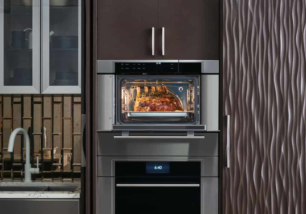 A modern kitchen showroom features a wall oven baking a seasoned roast, visible through the oven door, set beside a sink and textured cabinetry—a perfect display of luxury kitchen appliances.