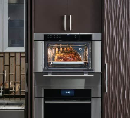 A modern kitchen showroom features a wall oven baking a seasoned roast, visible through the oven door, set beside a sink and textured cabinetry—a perfect display of luxury kitchen appliances.