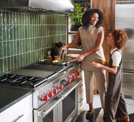 A woman and a girl stand in a modern kitchen, cooking together at a stove with green tiled backsplash and sleek Sub Zero appliance, resembling a stylish kitchen showroom.