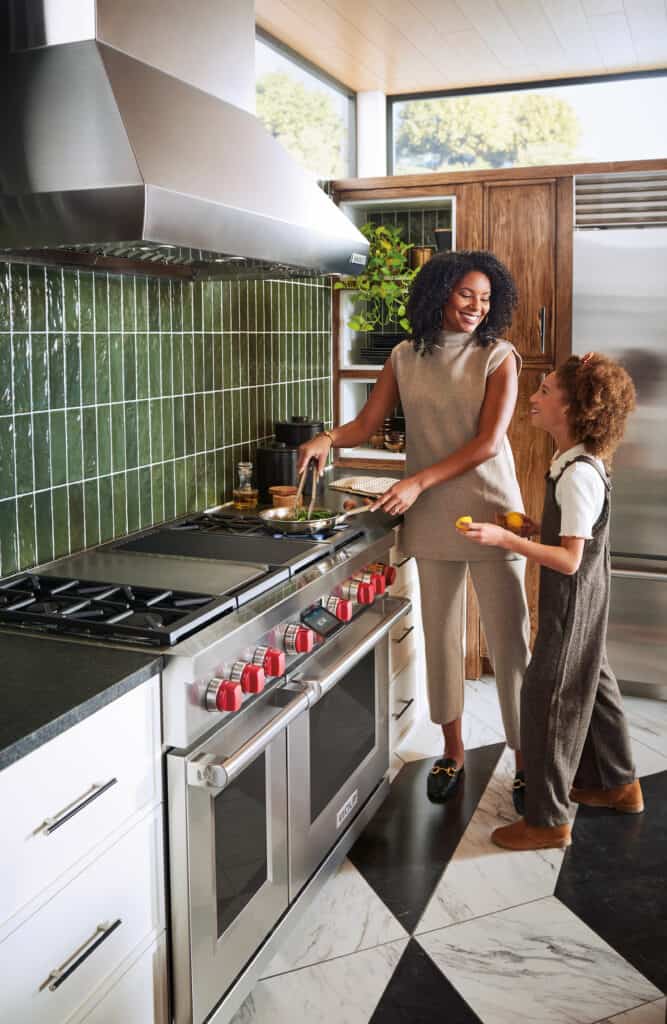 A woman and a girl stand in a modern kitchen, cooking together at a stove with green tiled backsplash and sleek Sub Zero appliance, resembling a stylish kitchen showroom.