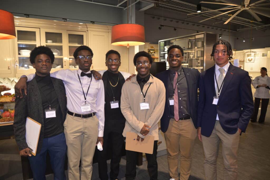Six young men wearing business attire and name badges stand together indoors, posing for a group photo in a well-lit kitchen showroom with luxury kitchen appliances and stylish displays in the background.