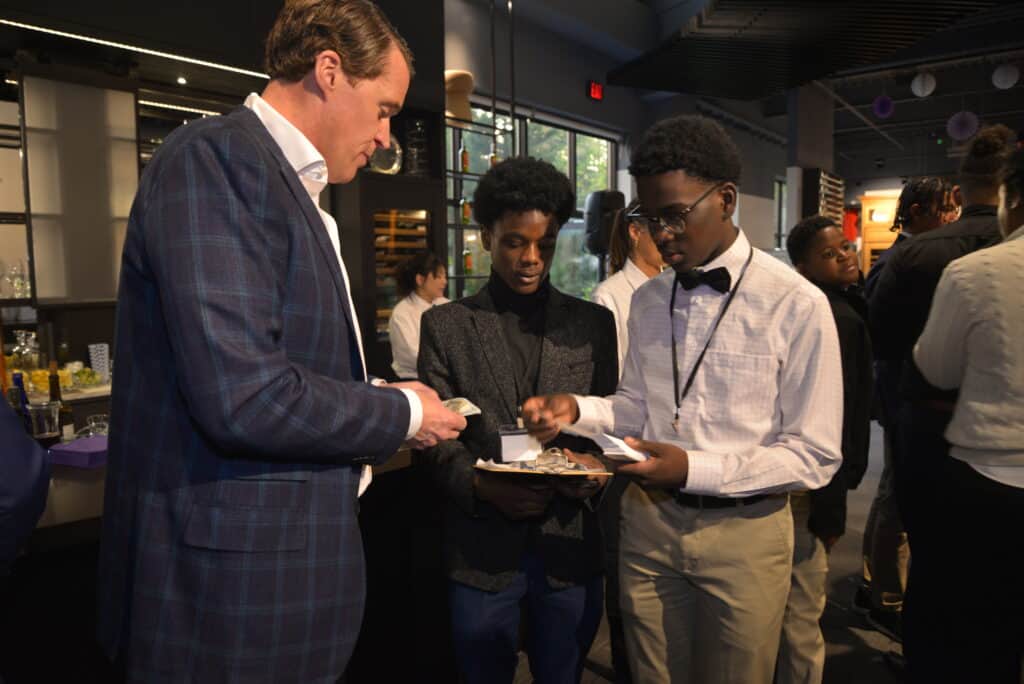 A man hands money to two young men holding a tray with cash and coins at a gathering in a modern appliance showroom.