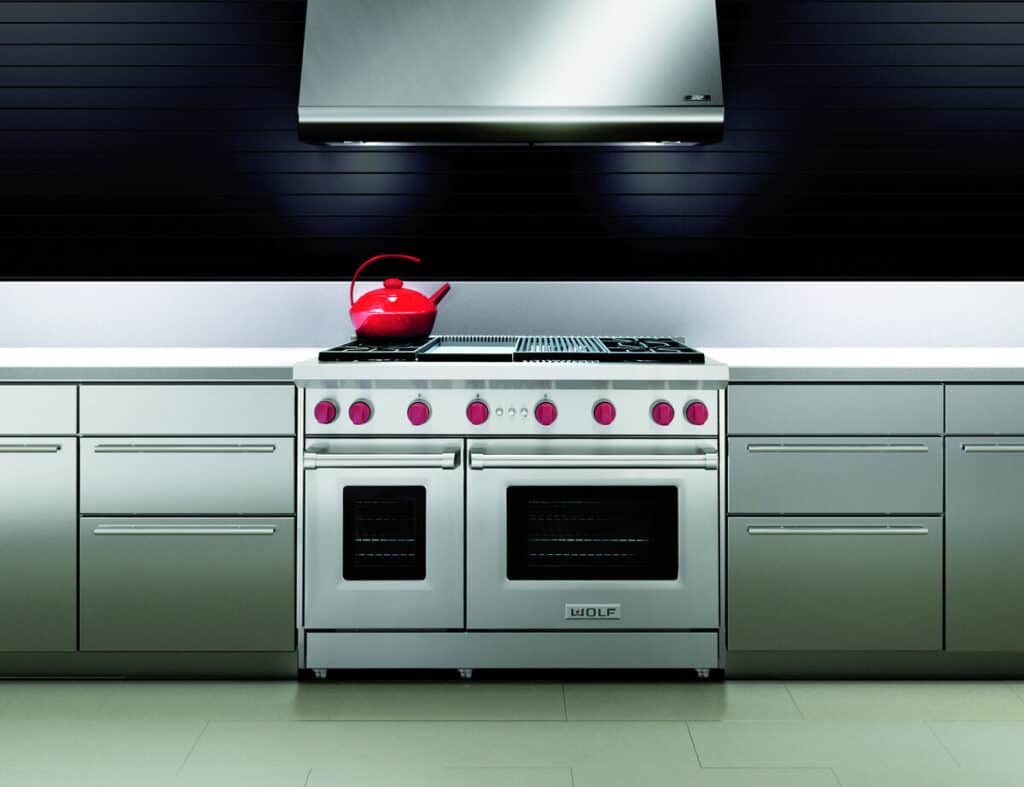 A stainless steel oven and range with red knobs, double doors, and a red kettle on top, set in a modern kitchen with gray cabinets and a metallic range hood, perfect for any luxury kitchen appliance showroom.