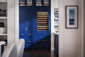 A built-in wine fridge, a luxury kitchen appliance with wooden shelves, is seamlessly integrated into blue cabinetry, next to a hallway with shelving and framed art on the wall.
