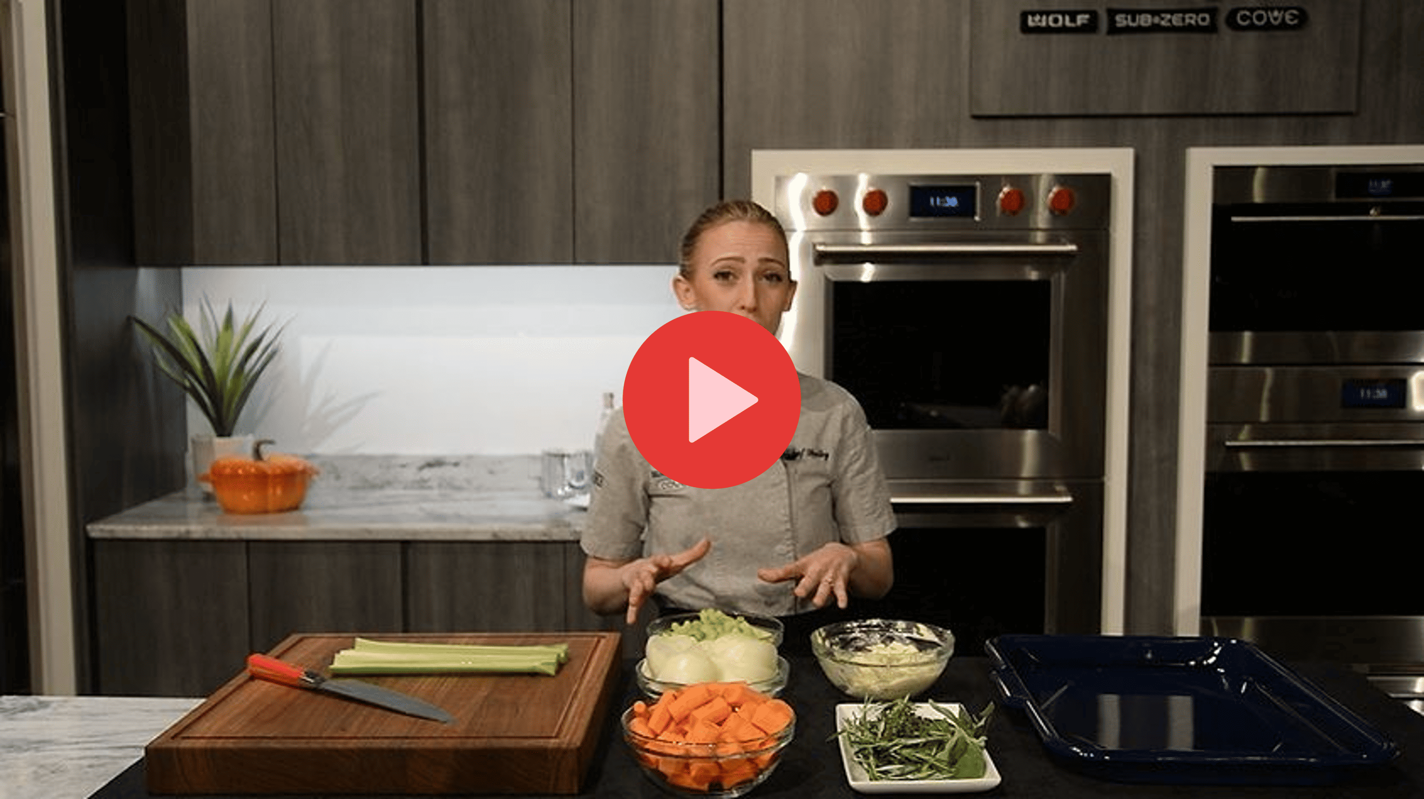 A chef stands in a modern kitchen behind a counter with chopped vegetables, a cutting board, and a knife, ready to demonstrate a recipe using luxury kitchen appliances. A play button icon is overlaid on the image.