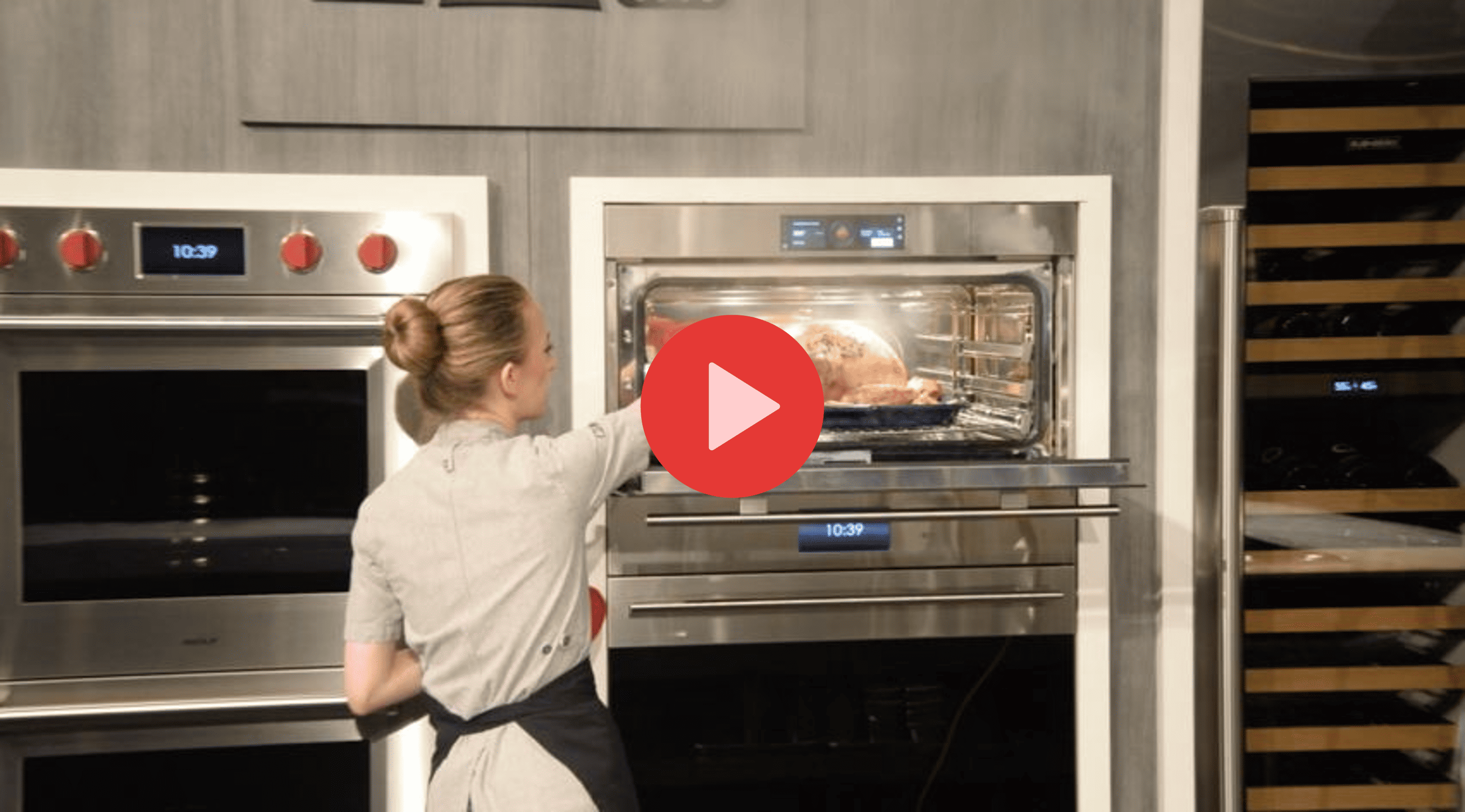 In a kitchen showroom, a woman in a gray uniform places a tray with food into a stainless steel oven, showcasing the latest sub zero appliance in the modern space.