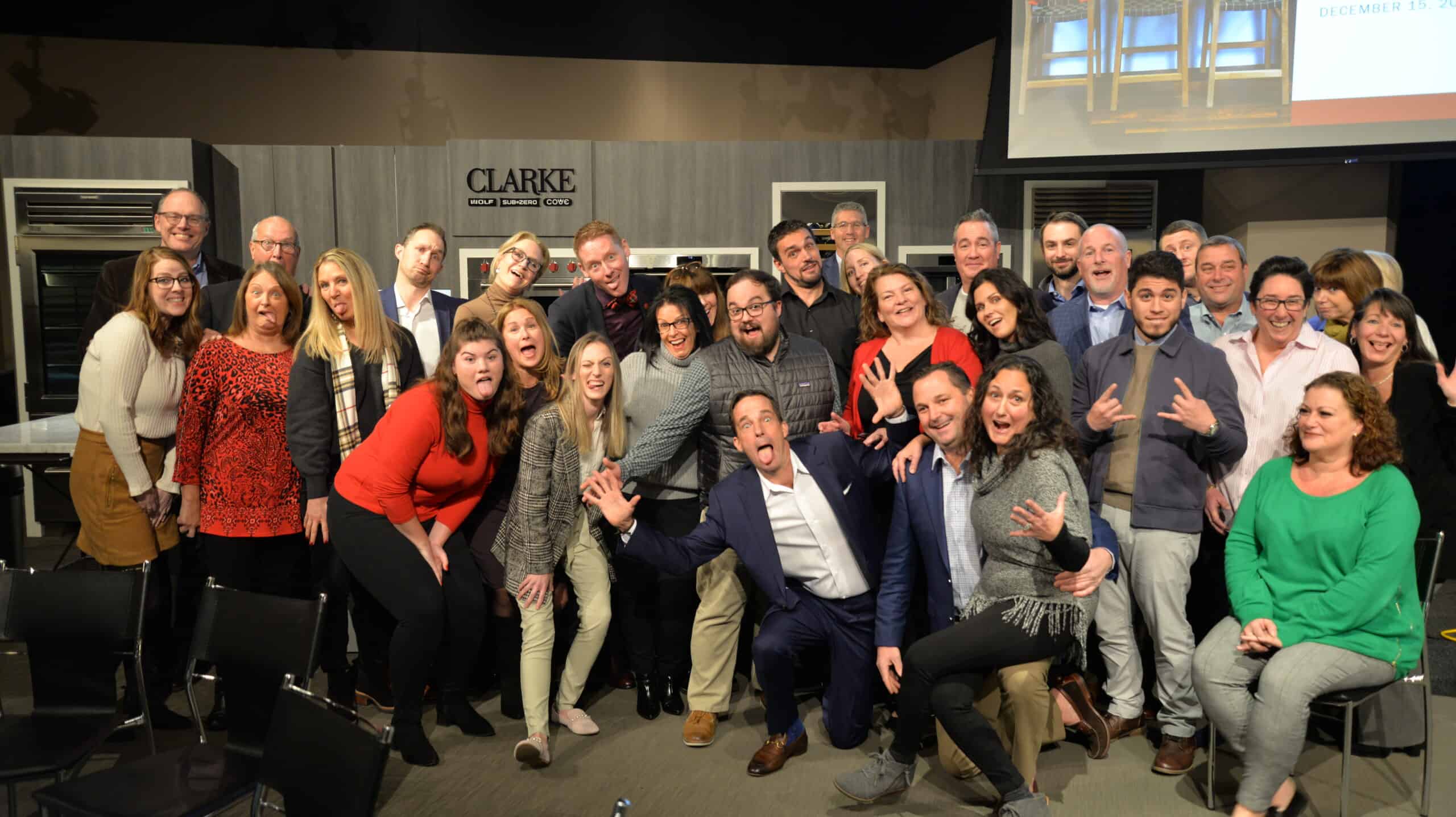 A group of adults pose together indoors, many smiling and making playful gestures, in front of a kitchen showroom display with "CLARKE" signage.