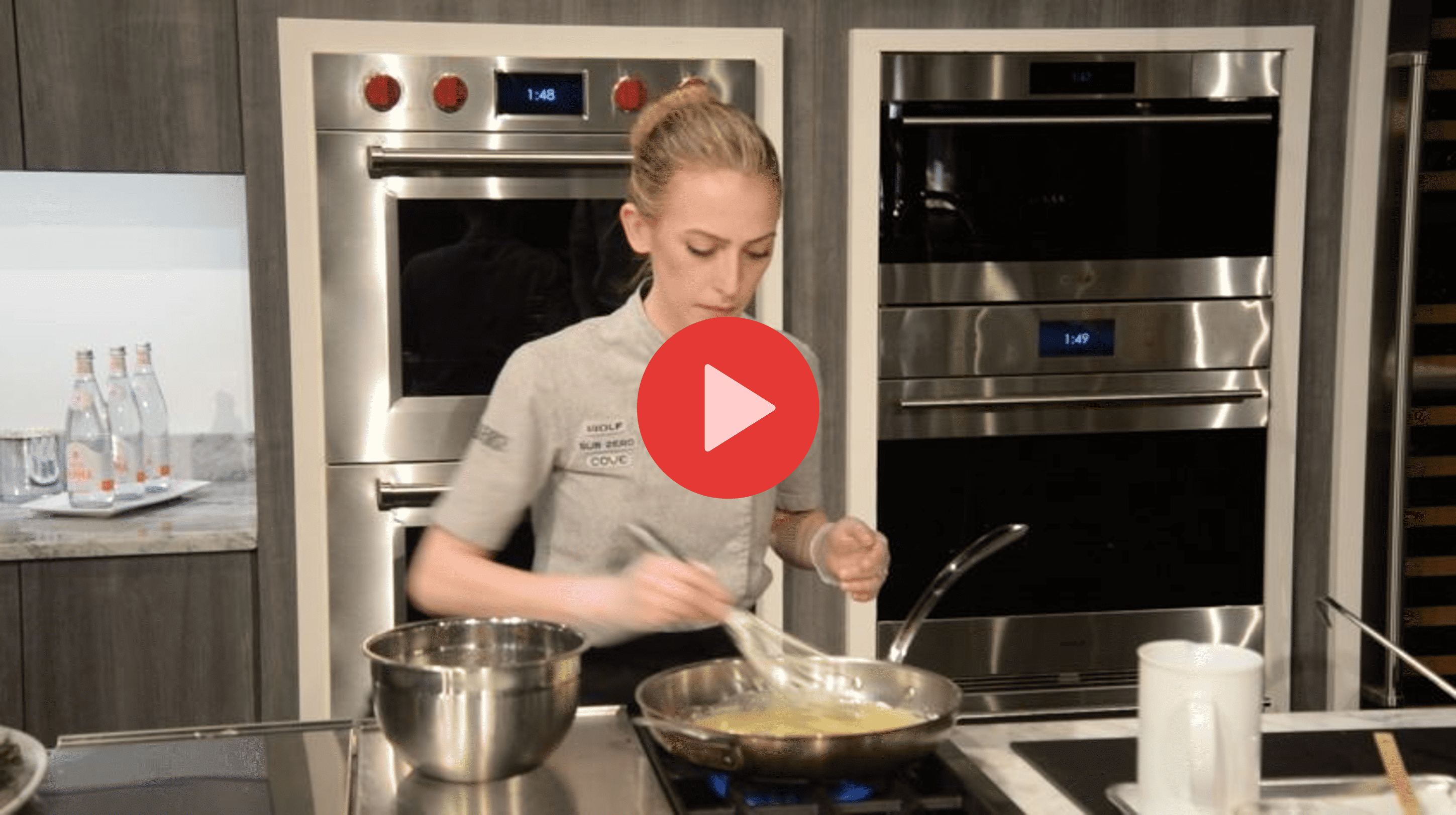 A woman in a kitchen is whisking ingredients in a pan on a stovetop, with metal mixing bowls and a sleek Wolf appliance visible among other appliances in the background.