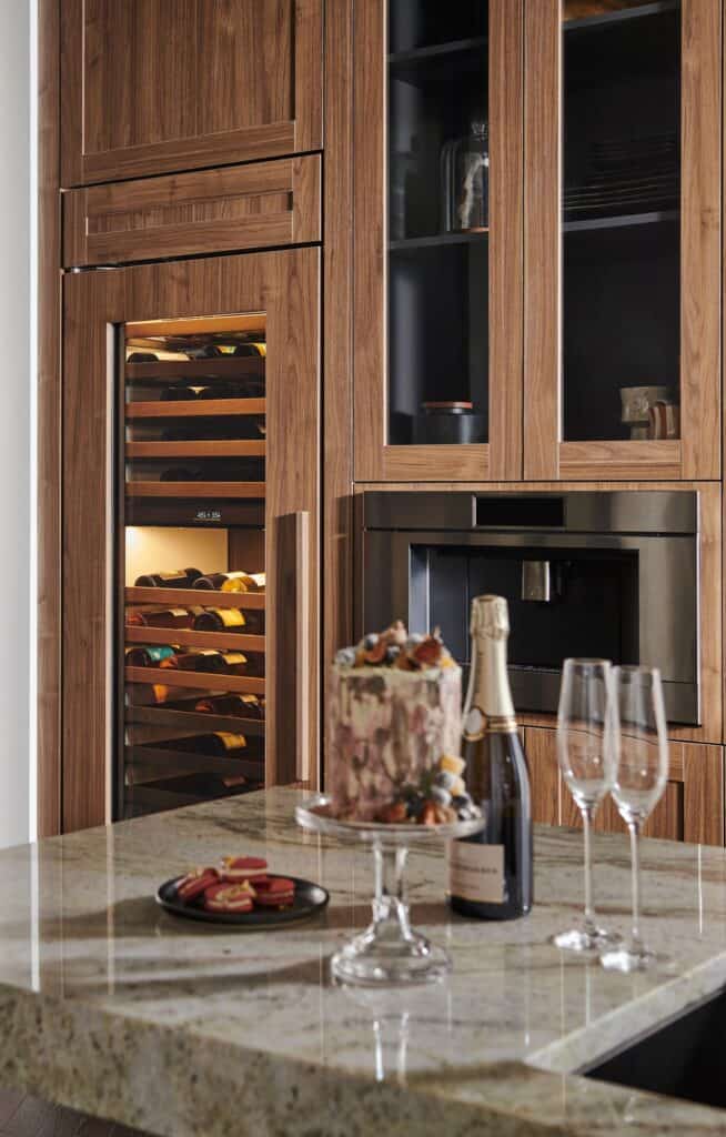 A modern kitchen features a built-in wine cooler with bottles, a cake on a stand, a Wolf appliance, champagne with two glasses, and desserts displayed on a marble countertop—perfect for showcasing luxury kitchen appliances.