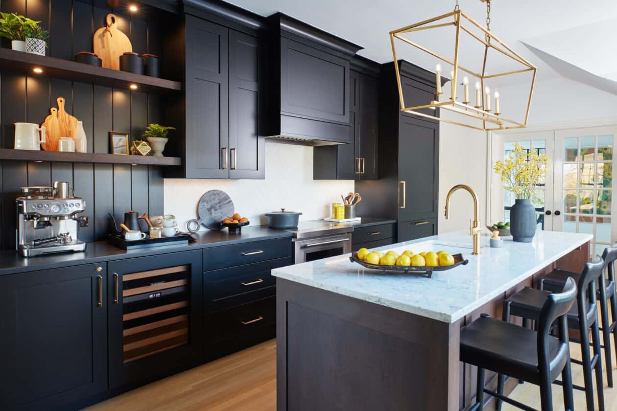 Modern kitchen showroom with dark cabinets, open shelves, marble island with stools, stainless steel appliances including a Wolf appliance, and a gold chandelier. Lemons and flowers decorate the countertop.