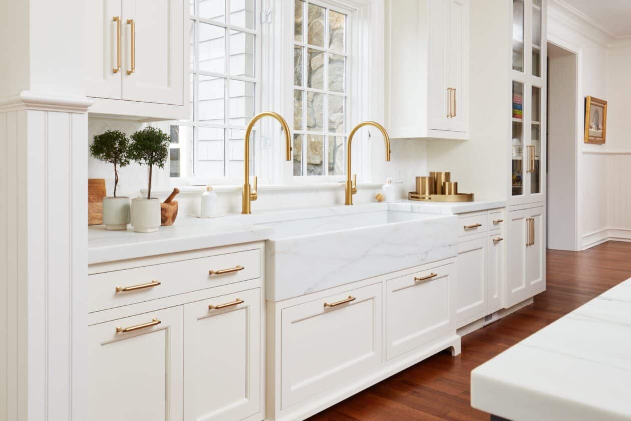 A modern kitchen with white cabinets, gold hardware, a marble countertop, two gold faucets, large windows above the sink, and a sleek Sub-Zero appliance that adds a touch of sophistication.