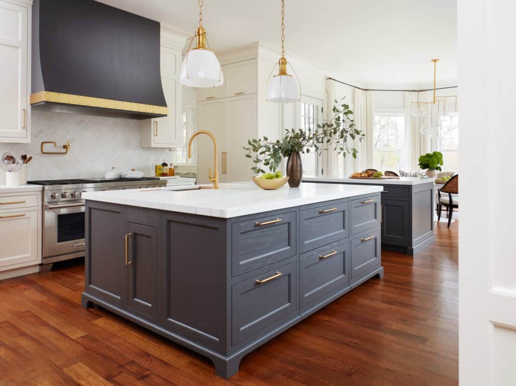 Modern kitchen with a large gray island, white countertops, gold fixtures, pendant lights, wood floors, and luxury kitchen appliances. A Sub Zero appliance and Wolf appliance complete the space; green apples and a vase with branches sit on the island.