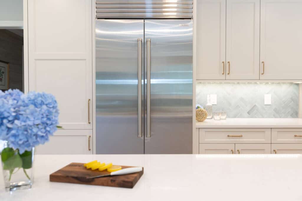 Modern kitchen showroom with a large stainless steel Sub-Zero appliance refrigerator, light cabinets, a vase of blue flowers, and a cutting board with a knife and sliced yellow bell pepper on the white counter.