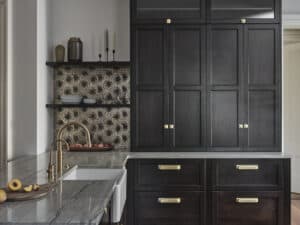 Modern kitchen with dark wood cabinets, brass hardware, a marble countertop, farmhouse sink, and geometric patterned backsplash. Open shelves hold dishes and decor, while a Wolf appliance adds a touch of elegance inspired by a kitchen showroom.