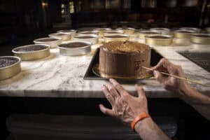 A person smooths chocolate frosting on a cake with a skewer on a marble countertop, surrounded by cake pans filled with batter, in a kitchen featuring a Sub Zero appliance—a true luxury kitchen appliance.