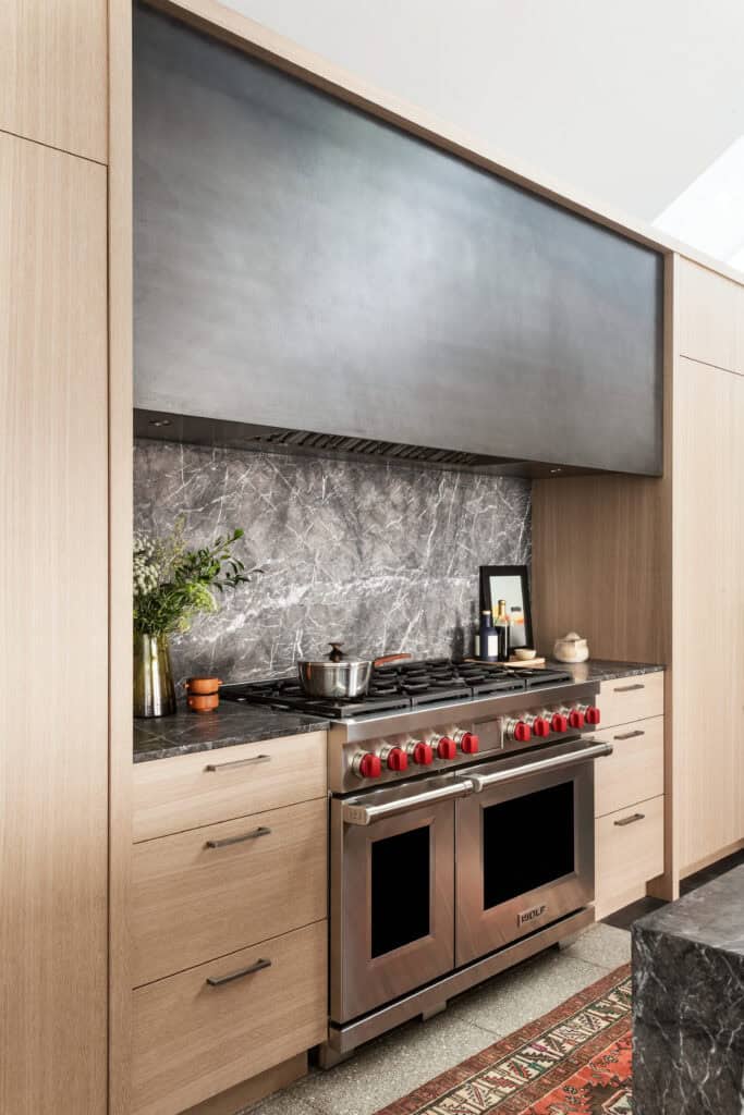 Modern kitchen with a stainless steel Wolf range, Sub-Zero appliance, marble backsplash, wooden cabinets, and kitchen items on the counter, including a vase with greenery—reminiscent of a luxury kitchen appliance showroom.