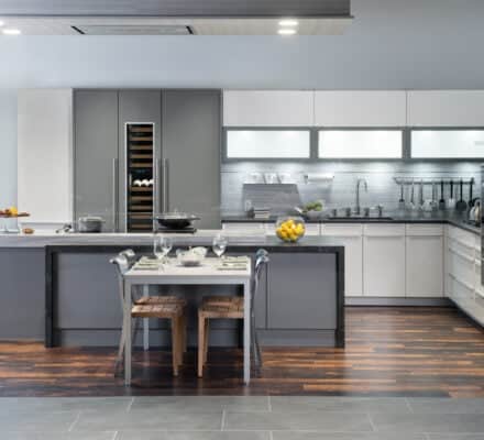Modern kitchen showroom with gray and white cabinets, a central island dining setup, wooden chairs, and luxury kitchen appliances including a Wolf appliance on dark wood and gray tile flooring.