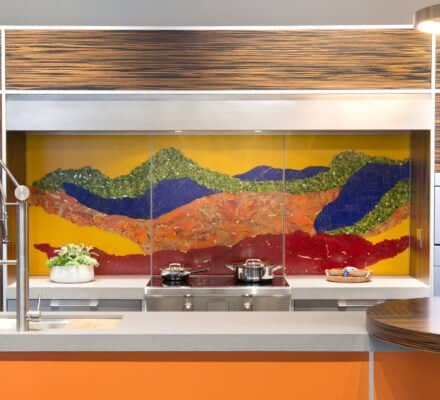 Modern kitchen showroom featuring a colorful mountain-themed backsplash, sleek luxury kitchen appliances, a built-in stovetop, and a potted plant on the counter.