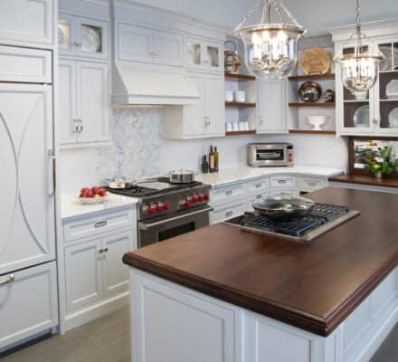 Modern kitchen showroom featuring white cabinets, a dark wood island countertop, stainless steel appliances including Sub-Zero and Wolf appliances, pendant lights, and open shelving displaying plates and decorative items.
