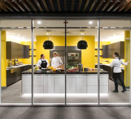 Three chefs work in a modern kitchen showroom with yellow walls; two talk near a counter with food, while one walks past quickly to the right, showcasing the latest Wolf appliance.
