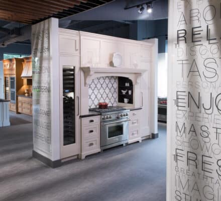 Modern kitchen showroom with white cabinetry, stainless steel appliances including a Sub Zero appliance, decorative backsplash, and pillars featuring large, embossed words.