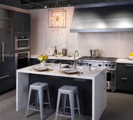 Modern kitchen with dark cabinets, a large stainless steel stove, Sub Zero appliance, built-in features, a white island with two metal stools, and decorative accents like flowers and a bowl of lemons—just like in a top kitchen showroom.