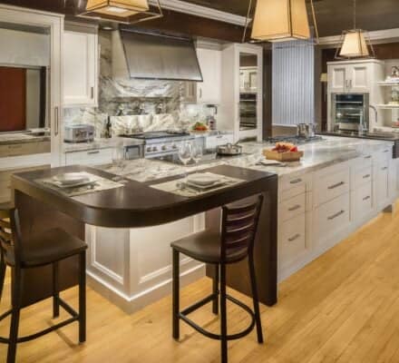 Modern kitchen with white cabinets, marble countertops, stainless steel appliances including a Wolf appliance, and a large island with two chairs and place settings. Wooden floor and pendant lights overhead create a true kitchen showroom feel.